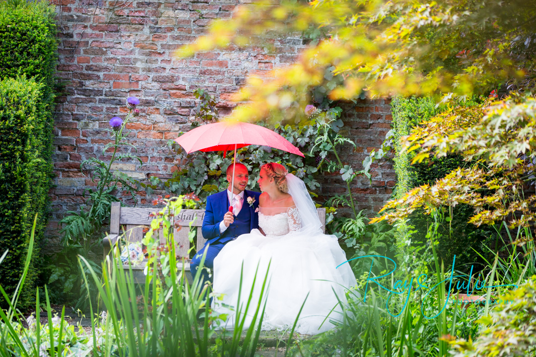 Bride and groom sat on a bench taking shelter from the rain under a love heart shaped umbrella, in the stunning grounds at Saltmarshe Hall.