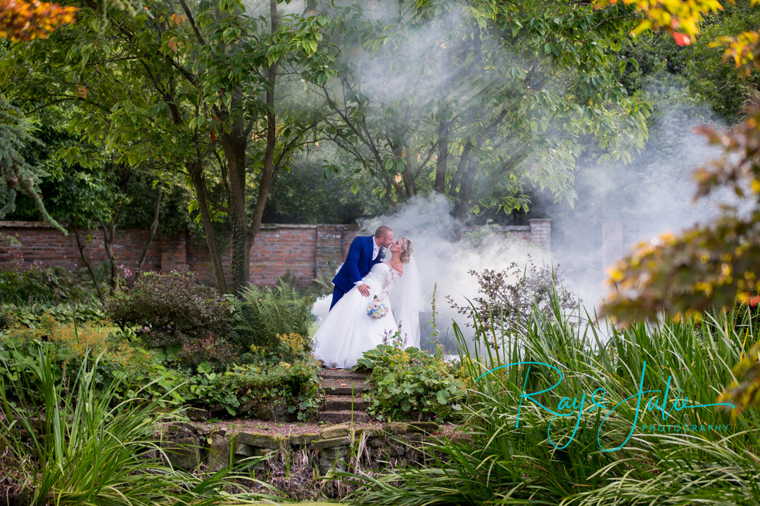 Stunning photograph of bride and groom bent over about to kiss in the grounds at Saltmarshe Hall. Smoke behind with flowers by All Occasions of Howden.