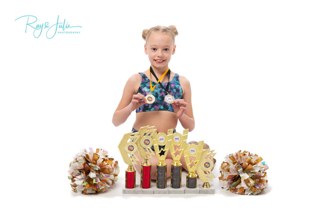 Young girl in a dance outfit holding medals, standing behind a collection of trophies with pom-poms on either side.