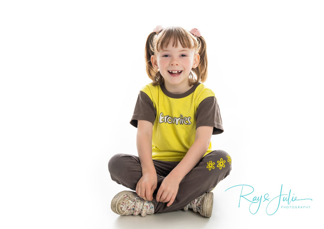 Young girl sitting cross-legged, smiling, wearing a yellow and brown "Brownies" shirt with pigtails against a white background.