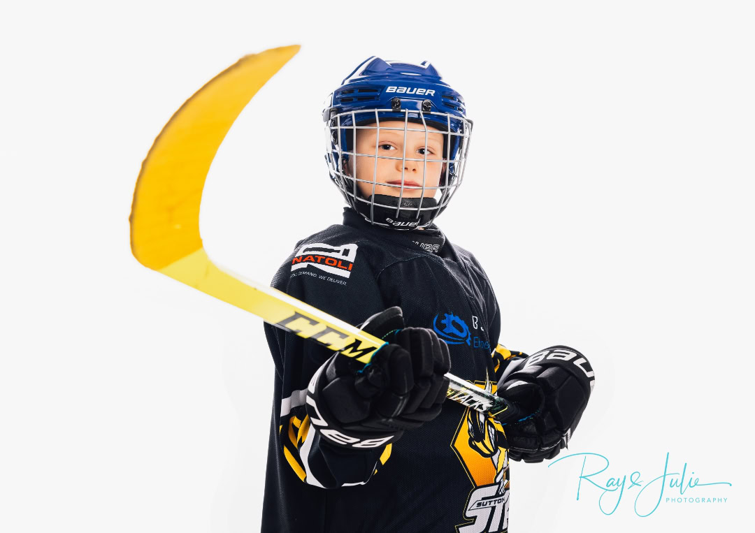 A young ice hockey player in a blue helmet and black uniform, holding a hockey stick with a yellow blade, poses against a white background.