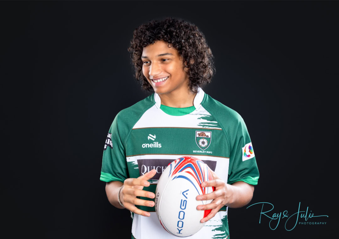 A person with curly hair smiles while holding a rugby ball. They are wearing a green and white rugby jersey with various logos.