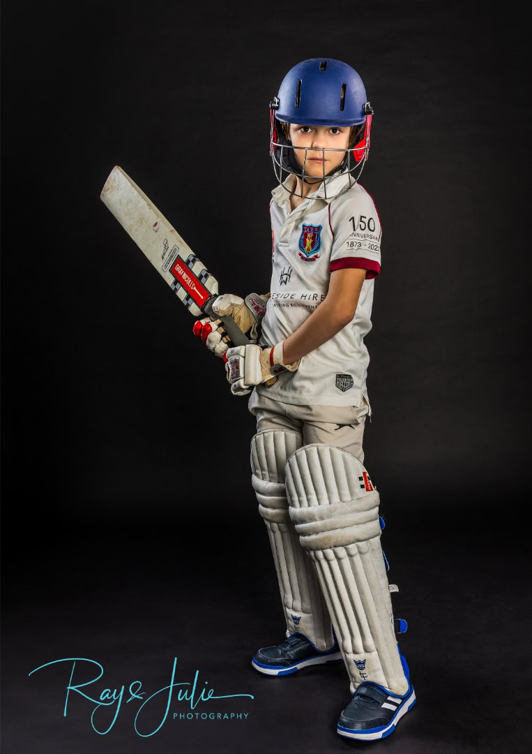 A child dressed in full cricket gear, including a helmet, pads, and gloves, holding a bat, stands against a dark background.