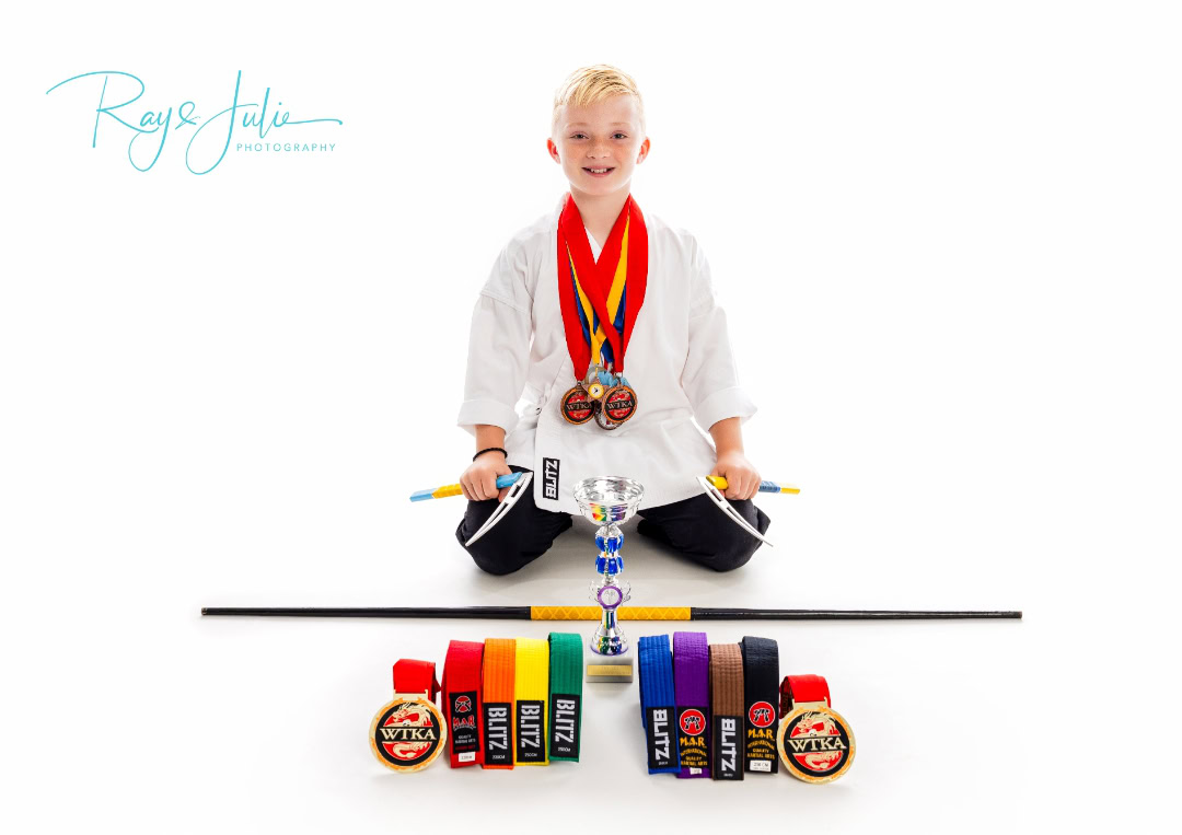 Young boy in a martial arts uniform sitting with a trophy, medals around his neck, and various colored belts displayed in front of him.