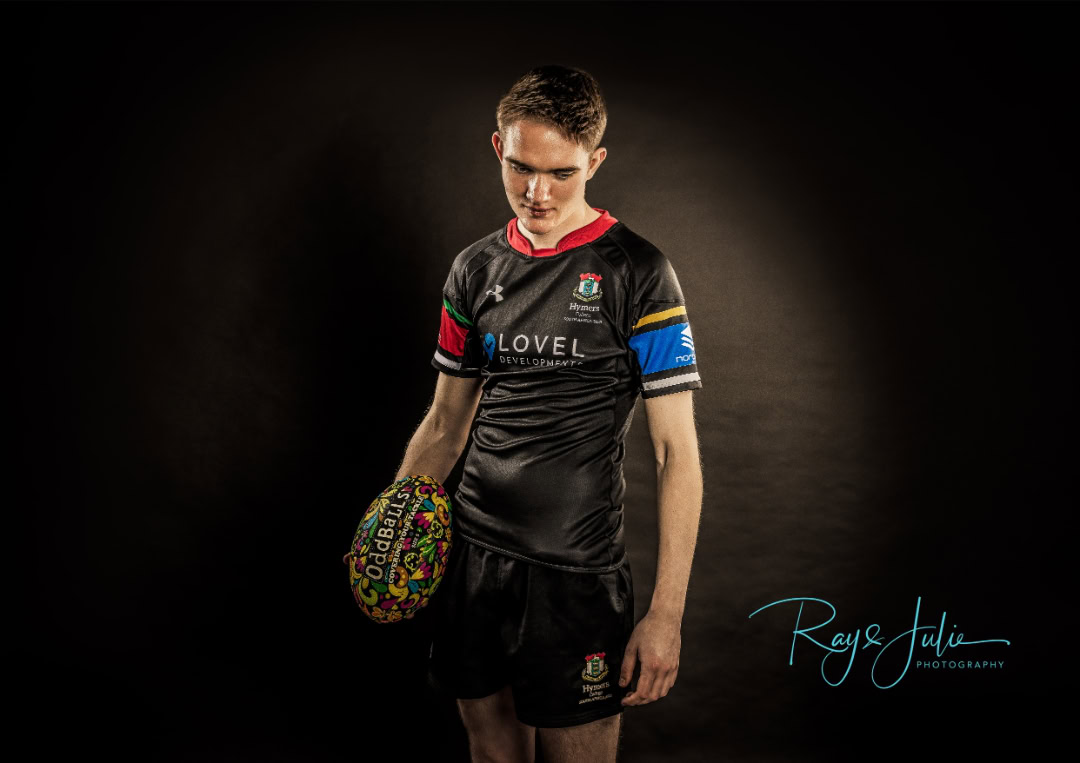 A rugby player in a black uniform stands holding a colorful rugby ball against a dark background.