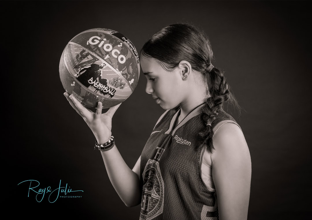 A woman holding a patterned basketball near her forehead, wearing a sleeveless sports jersey.