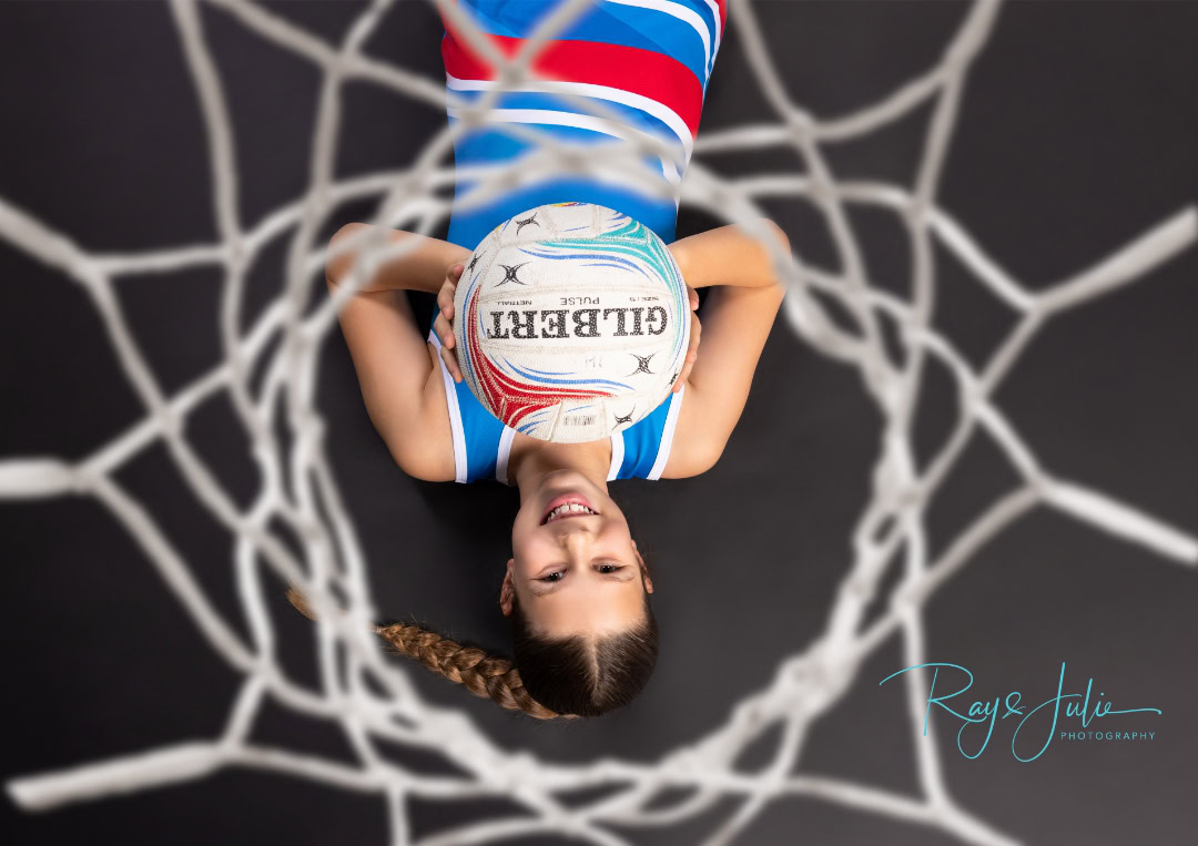 Girl lying on ground, smiling, with a braided hair and a netball resting on her chest, viewed through a netball hoop from above.