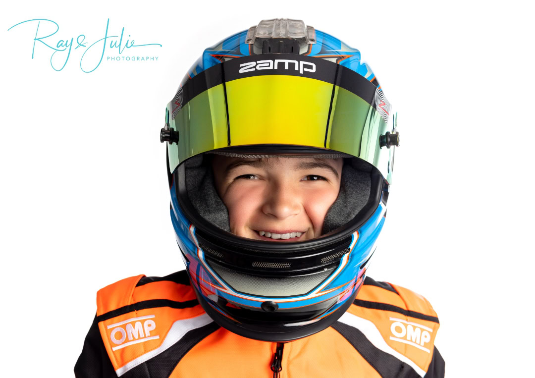 Child wearing a blue racing helmet and orange racing suit, smiling at the camera.