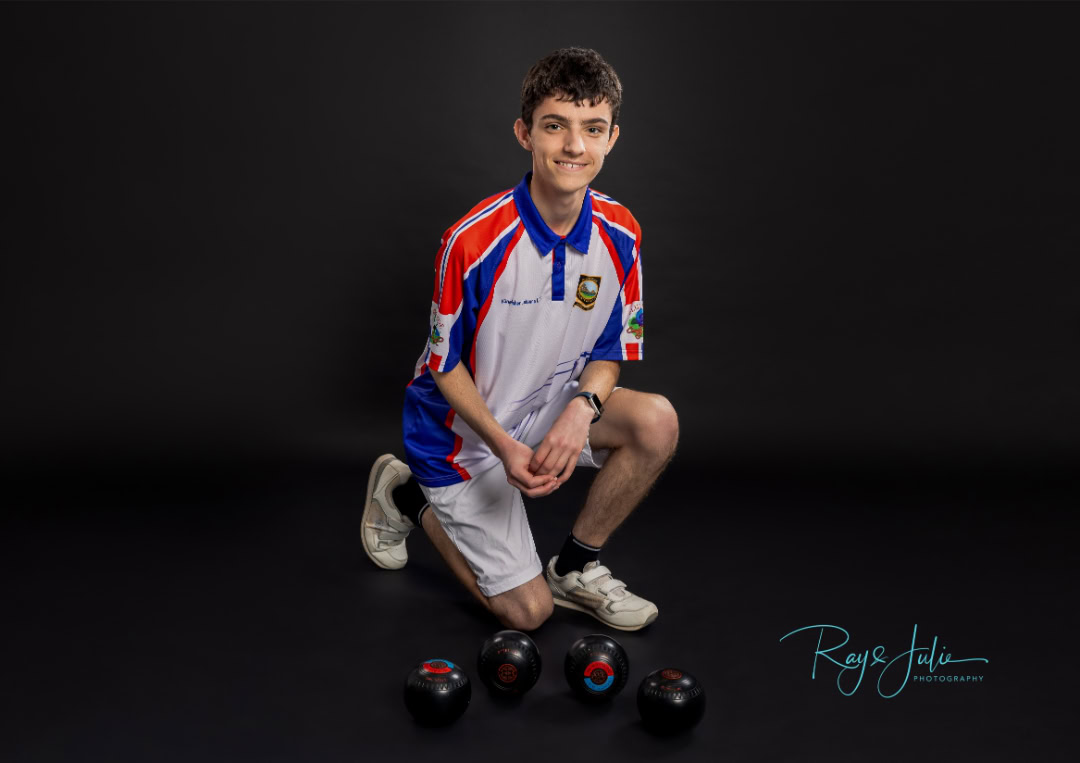 A young man in a sports uniform kneels on one knee in front of several bowling balls on a black background.