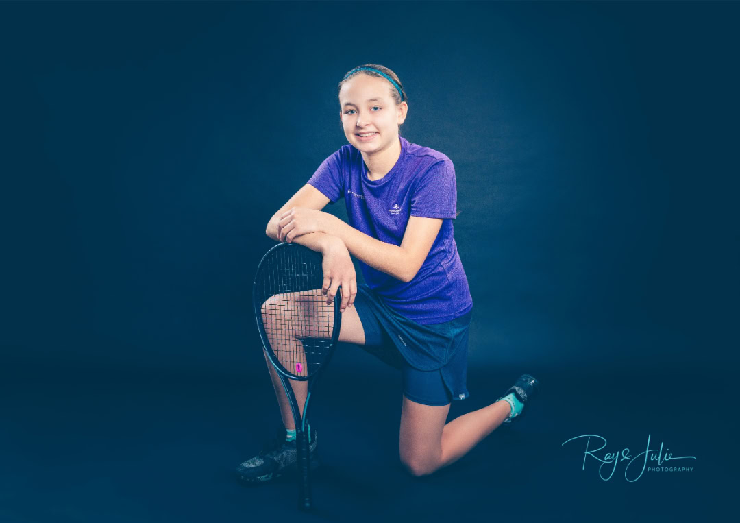 Person kneeling on one knee, holding a tennis racket. Wearing a purple shirt and shorts, against a dark background.