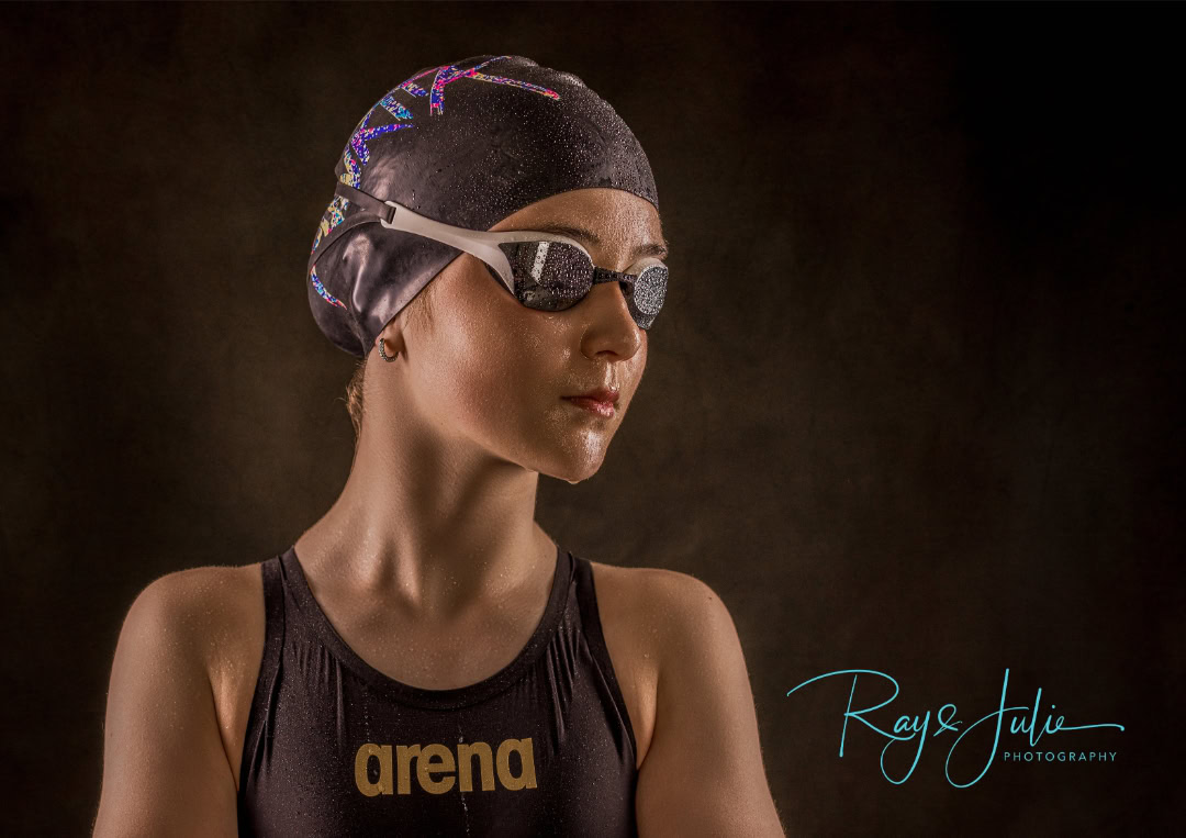 A swimmer wearing a black cap, goggles, and a swimsuit with the brand "arena" looks to the side against a dark background.