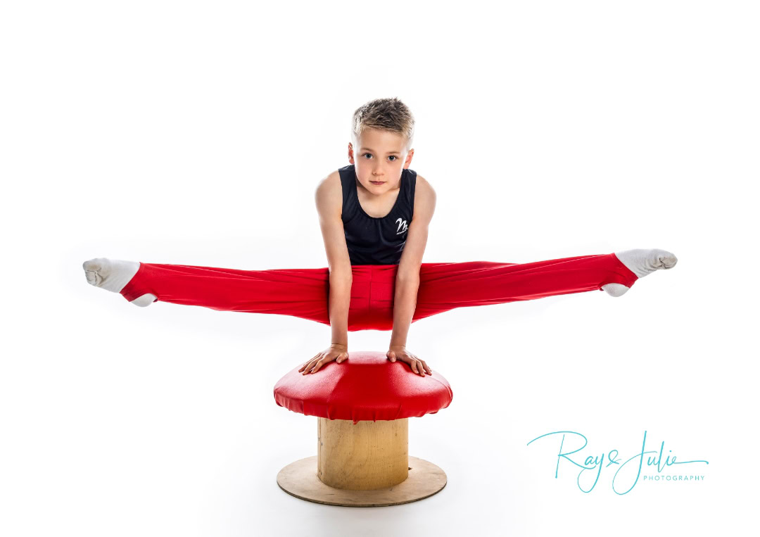 A young gymnast performs a straddle press handstand on a mushroom, wearing a black tank top and red pants against a white background.