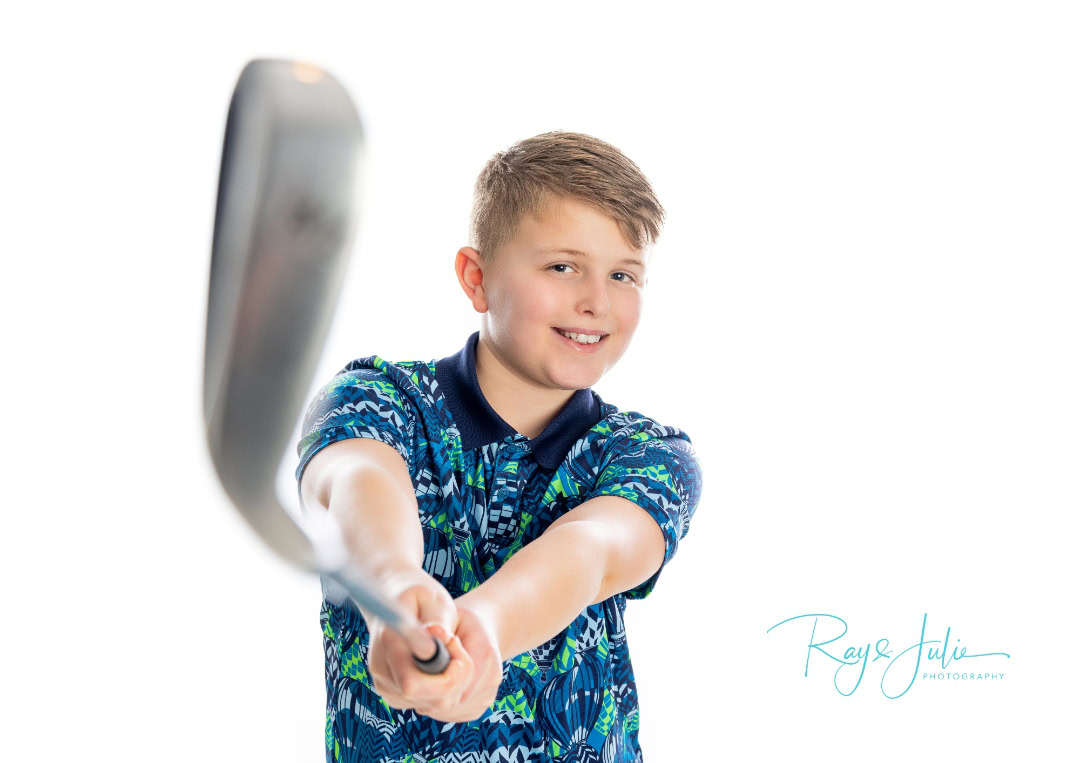 Boy smiling and holding a golf club toward the camera, wearing a patterned shirt. White background.