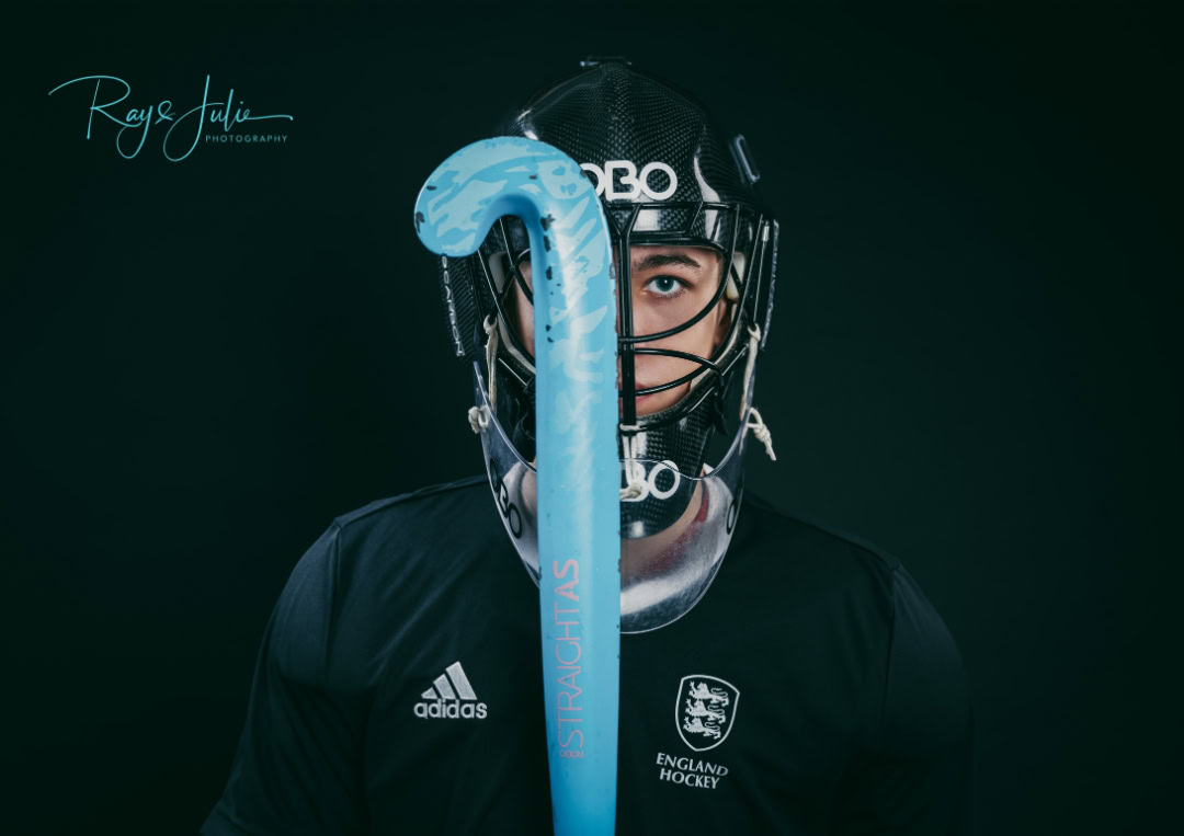 A field hockey player wearing a helmet and black uniform holds a blue hockey stick vertically in front of their face.