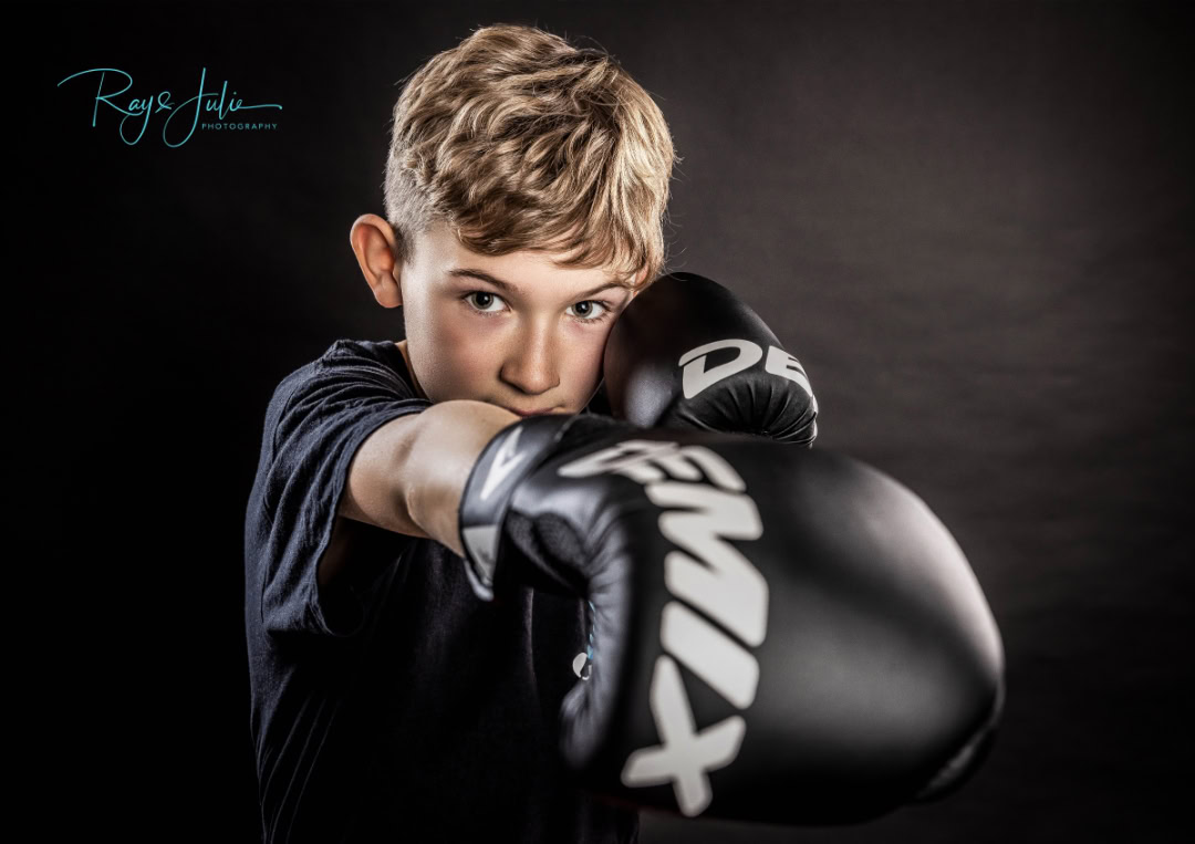 A young person wearing boxing gloves poses with a focused expression against a dark background.