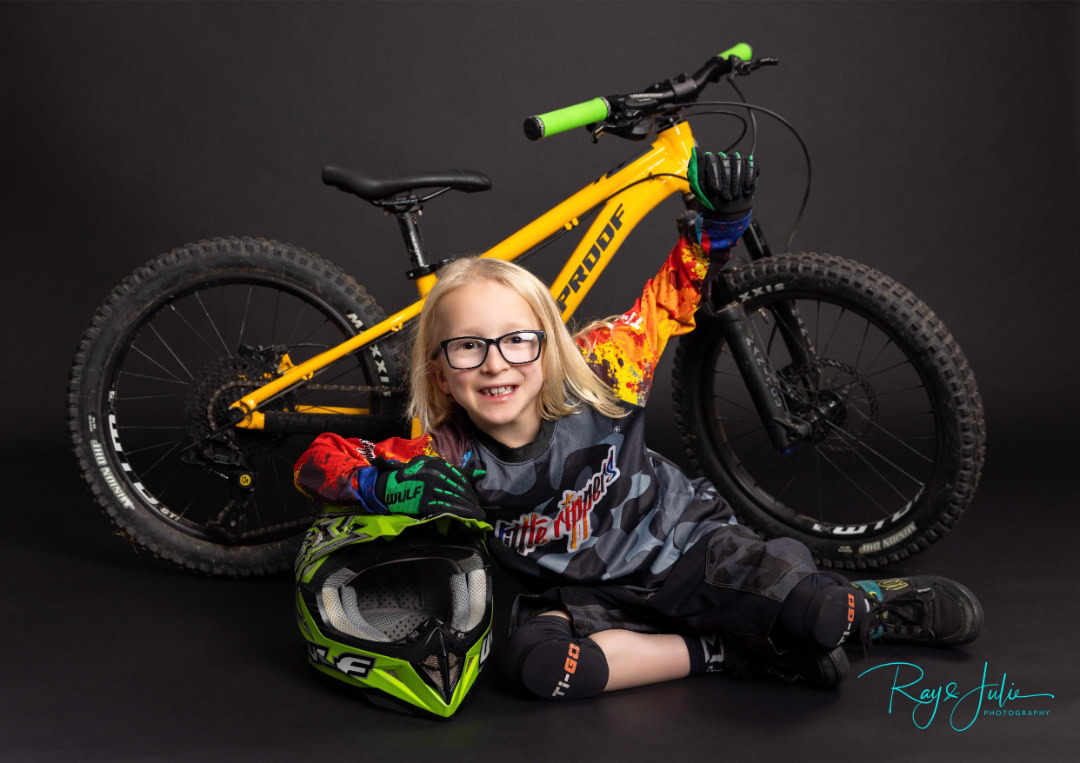 A child wearing biking gear sits on the floor with a neon green helmet, posing in front of a yellow mountain bike against a black background.