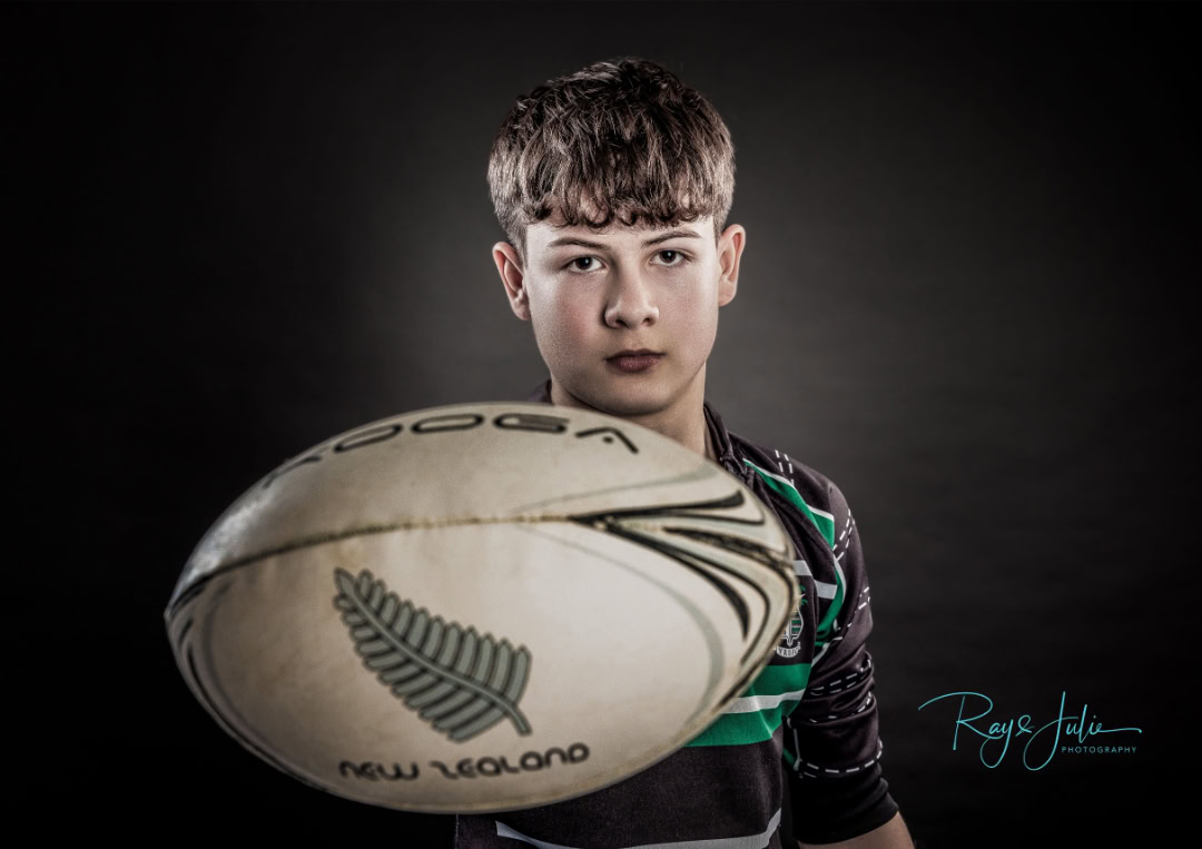 A boy holds a rugby ball with a New Zealand logo, wearing a striped rugby jersey, against a dark backdrop.